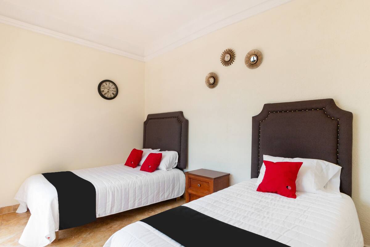 Habitación de hotel con dos camas, cada una con cabecero marrón, ropa de cama blanca y almohadas rojas. Entre las camas hay una mesita de noche de madera, un reloj de pared y tres apliques decorativos.