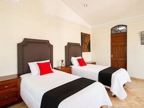 A hotel room with two beds, each with a headboard, white bedding, and a black accent. There is a wooden nightstand between the beds, a small table with a vase, and a wooden door with a window.