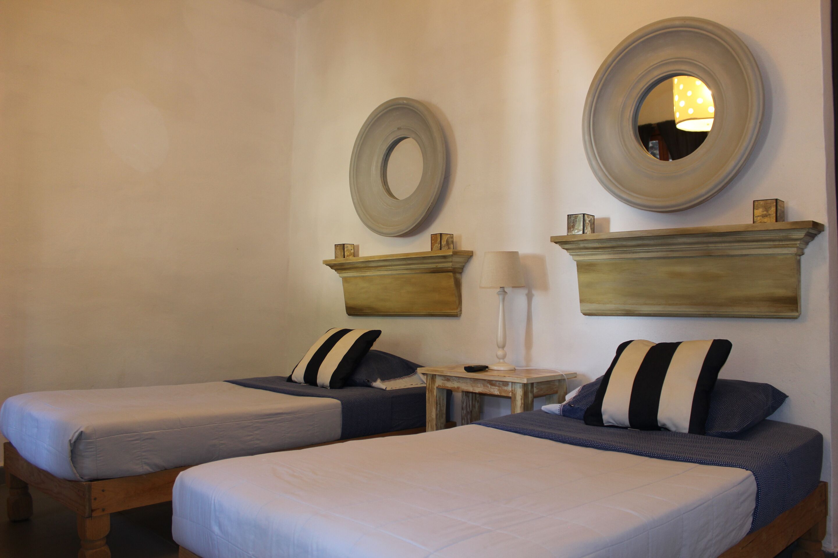 A room with two beds, each with a pillow and a bedside table with a lamp. There are wall-mounted mirrors and shelves above the beds.