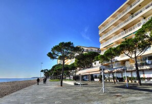 Un paseo marítimo empedrado con losetas de piedra, árboles y una playa al fondo.
