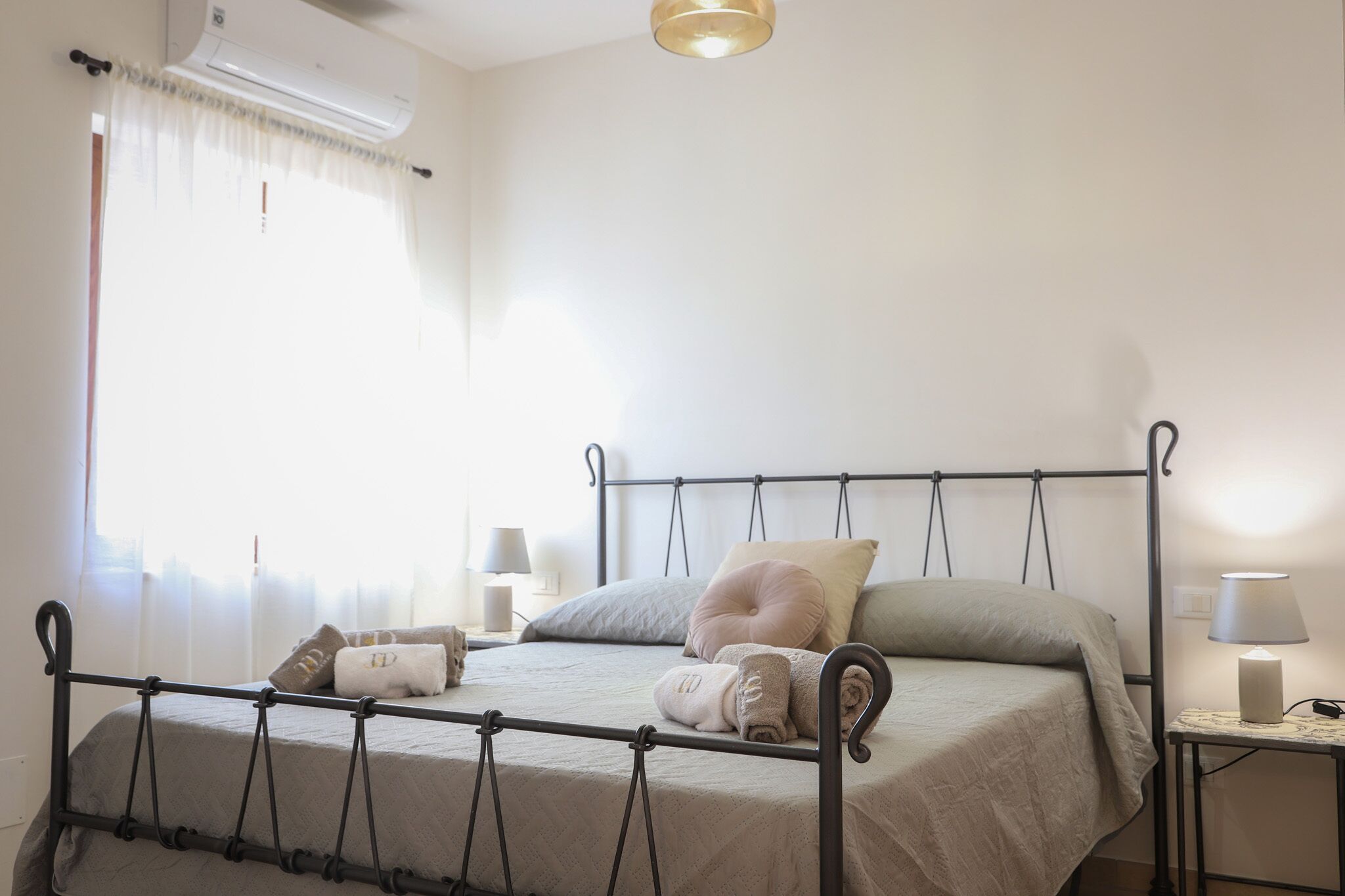 A neatly made bed with a metal frame, a bedside table with a lamp, and a window with sheer curtains.