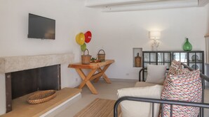 A living room with a fireplace, a television mounted on the wall, a dining table with baskets and fruits, a sofa with patterned cushions, and a cabinet with glass doors. - TD Trullo Roverella Charming Trullo with Pool (Castellana Grotte)