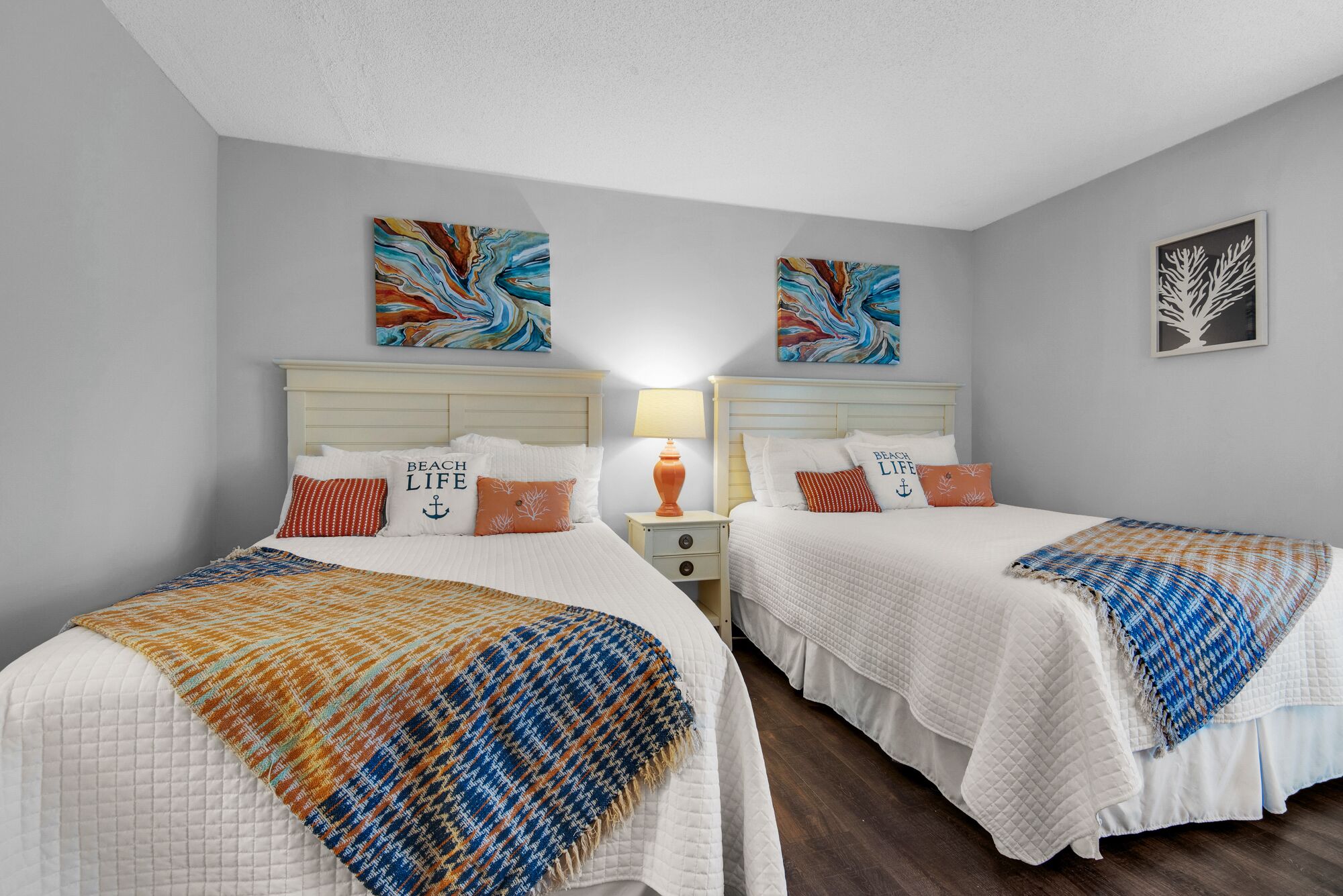 Une chambre d’hôtel avec deux lits, chacun recouvert d’une courtepointe blanche matelassée et agrémenté d’oreillers assortis. Deux œuvres d’art sont accrochées au mur au-dessus des lits, et une image encadrée se trouve sur le mur, à droite.