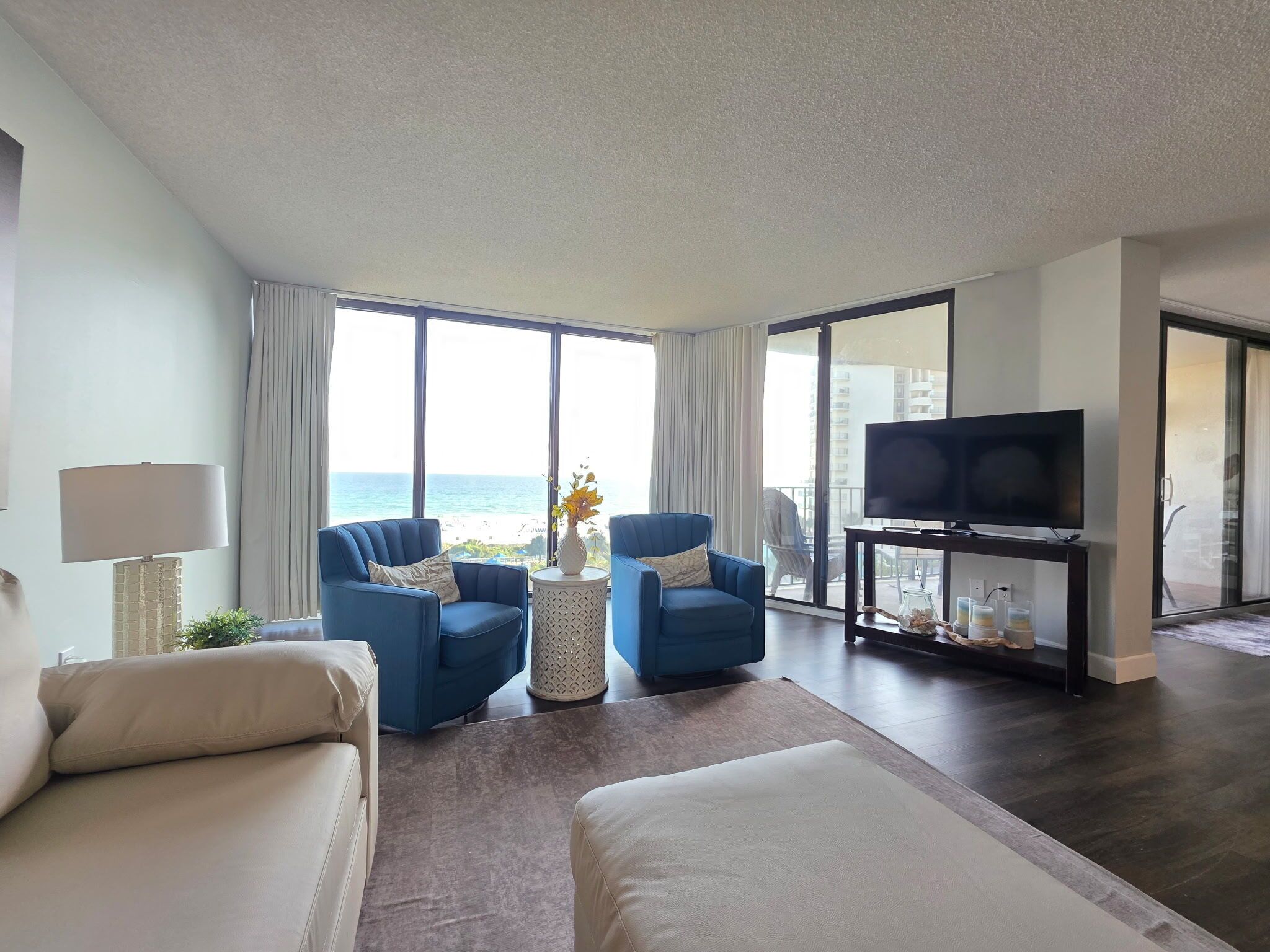 Een moderne hotelkamer met een groot raam met uitzicht op zee, twee blauwe fauteuils, een witte bank, een klein bijzettafeltje met een vaas, en een flatscreen televisie op een standaard.