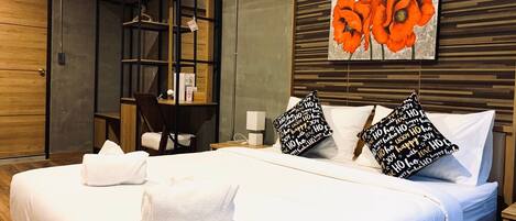 A hotel room with a large bed, a wooden desk, a bookshelf, and a painting of orange flowers on the wall.