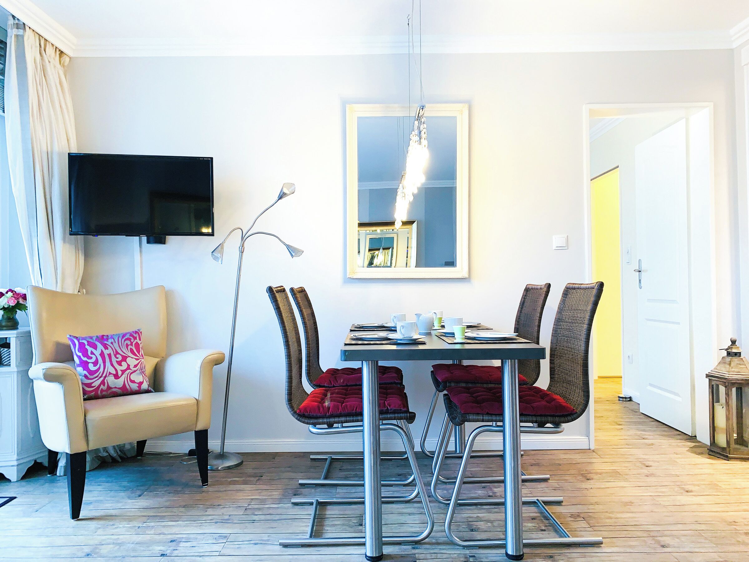 A modern dining area with a table set for four, surrounded by chairs with red cushions. A floor lamp and a flat-screen TV are mounted on the wall.