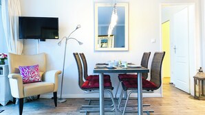 Ett modernt matområde med ett bord för fyra personer, omgivet av stolar med röda kuddar. En golvlampa och en platt-TV är monterade på väggen.