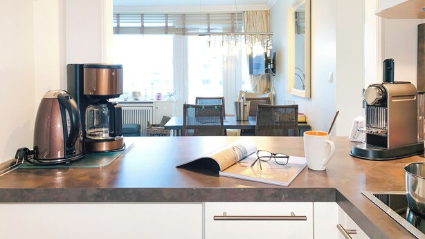 Ett modernt kök med en kaffebryggare, vattenkokare och espressomaskin. Här finns också en matplats med ett bord och stolar, samt ett fönster med gardiner.