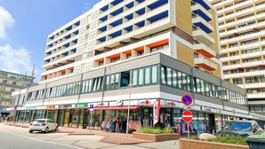 A multi-story building with a modern architectural design, featuring a ground-level retail space with visible storefronts and a parking area. - House by the Sea App. 21 (Westerland)