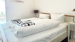 A neatly made bed with white bedding and striped towels, a bedside lamp, and a small table with a lamp. - House by the Sea App. 21 (Westerland)