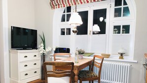 A dining area with a wooden table, two chairs, a television on a cabinet, and a window with shutters. - Lille Villa App Stars Westerland (Westerland)