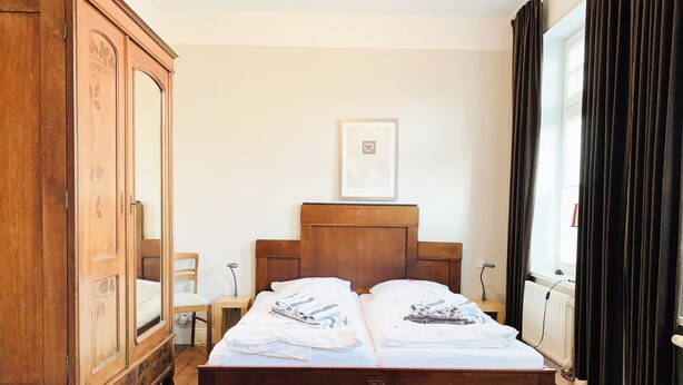 A bedroom with a wooden bed, a wooden wardrobe, a small bedside table, a chair, and a window with curtains. - Lille Villa App Stars Westerland (Westerland)