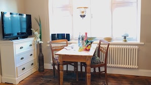 A dining area with a wooden table, chairs, a television on a white cabinet, a radiator, and a window with a view. - Lille Villa App Stars Westerland (Westerland)