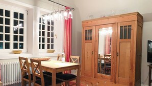 A dining area with a wooden table and chairs, a radiator, a large wooden wardrobe, and a window with white frames and a red curtain. - Lille Villa App Moon Westerland (Westerland)
