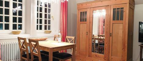A dining area with a wooden table and chairs, a radiator, a large wooden wardrobe, and a window with white frames and a red curtain.