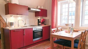 A compact kitchen with red cabinets, a built-in oven, and a dining table with chairs. - Lille Villa App Moon Westerland (Westerland)