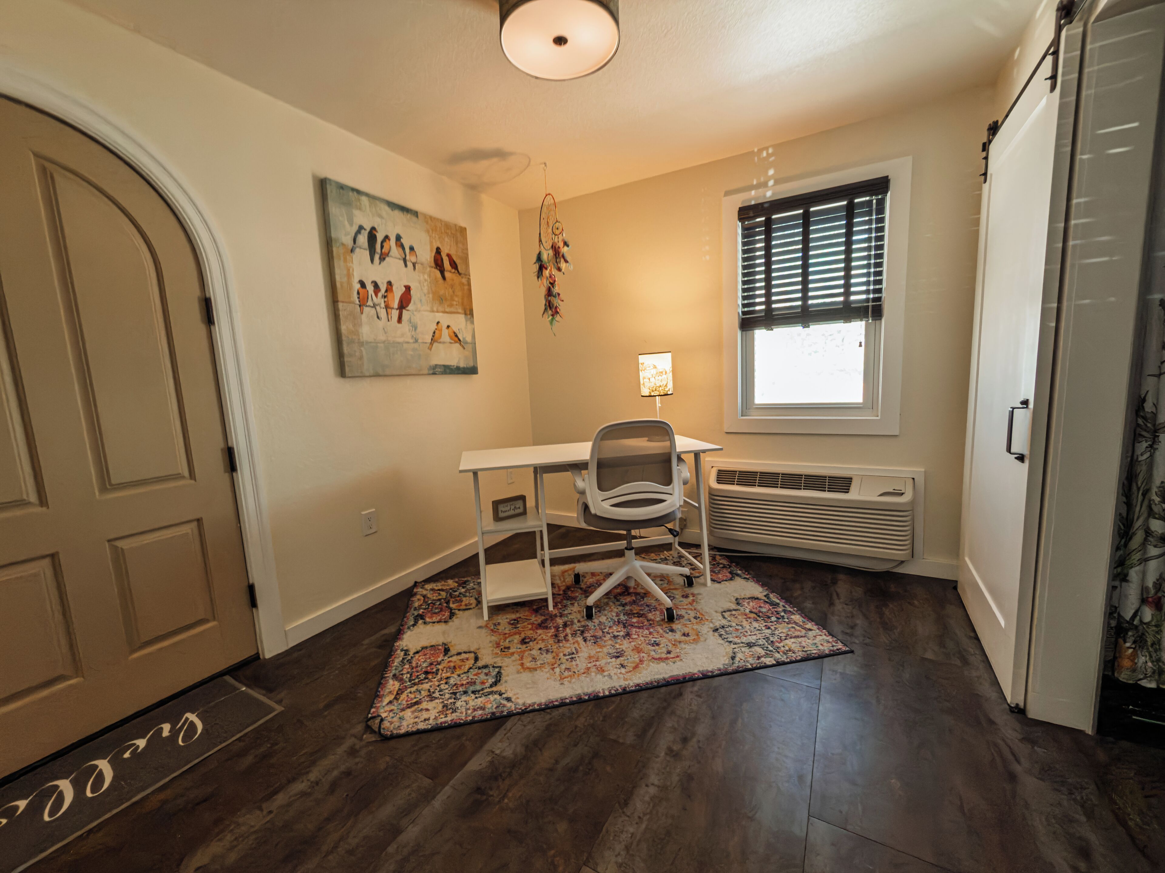 A room with a white desk, a chair, a patterned rug, a wall-mounted dreamcatcher, a window with blinds, and a radiator.