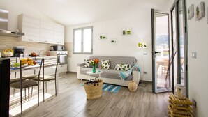 A modern kitchen with a dining area, a sofa, and a view through a glass door. - Flower house - Charming cottage 500 meters from the beach (Tertenia)