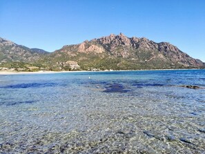 A beach with clear shallow waters, a mountain in the background, and a sandy shore.