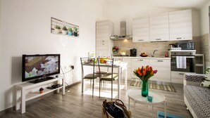 A modern living room with a flat-screen TV, a dining area with chairs, a small table with a vase of flowers, and a kitchen area with cabinets and appliances. - Flower house - Charming cottage 500 meters from the beach (Tertenia)