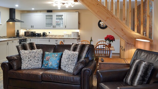 A cozy living room with a brown leather sofa, a wooden staircase, and a kitchen in the background.