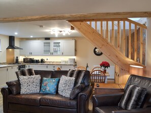 A cozy living room with a brown leather sofa, a wooden staircase, and a kitchen in the background.