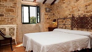 A room with a stone wall, a bed with a white bedspread, a wooden chair, and a window with a view of greenery. - STONE HOUSE WITH PORCH AND TERRACE  (Laxe)