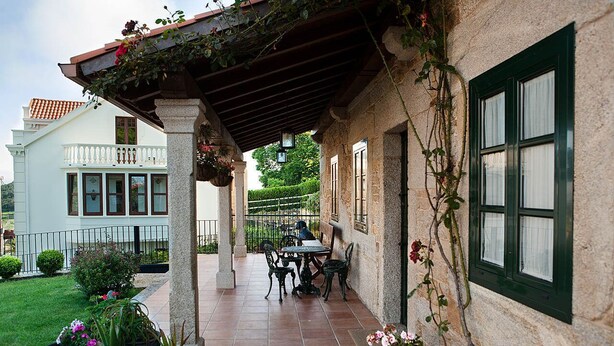 A covered patio with a table and chairs, potted plants, and a view of a white building with a balcony. - STONE HOUSE WITH PORCH AND TERRACE  (Laxe)