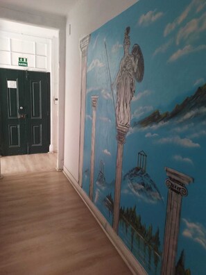 Un pasillo con un mural de arquitectura clásica, una puerta verde con el rótulo 'SAÍDA' y suelo de madera.