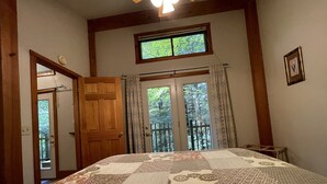 A bedroom with a large bed, a wooden door, a window with curtains, and a small table. - Hot Tub in the treetops with Timber Framed 2 bedroom cabin hidding in the woods (Victor)