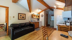 A cozy living room with a black leather sofa, wooden dining table, and a kitchen area with wooden beams and a white refrigerator. - Hot Tub in the treetops with Timber Framed 2 bedroom cabin hidding in the woods (Victor)