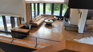 Ein geräumiges Wohnzimmer mit einer großen Eckcouch, einem Couchtisch und einem Kamin. Große Fenster bieten einen Blick ins Freie.