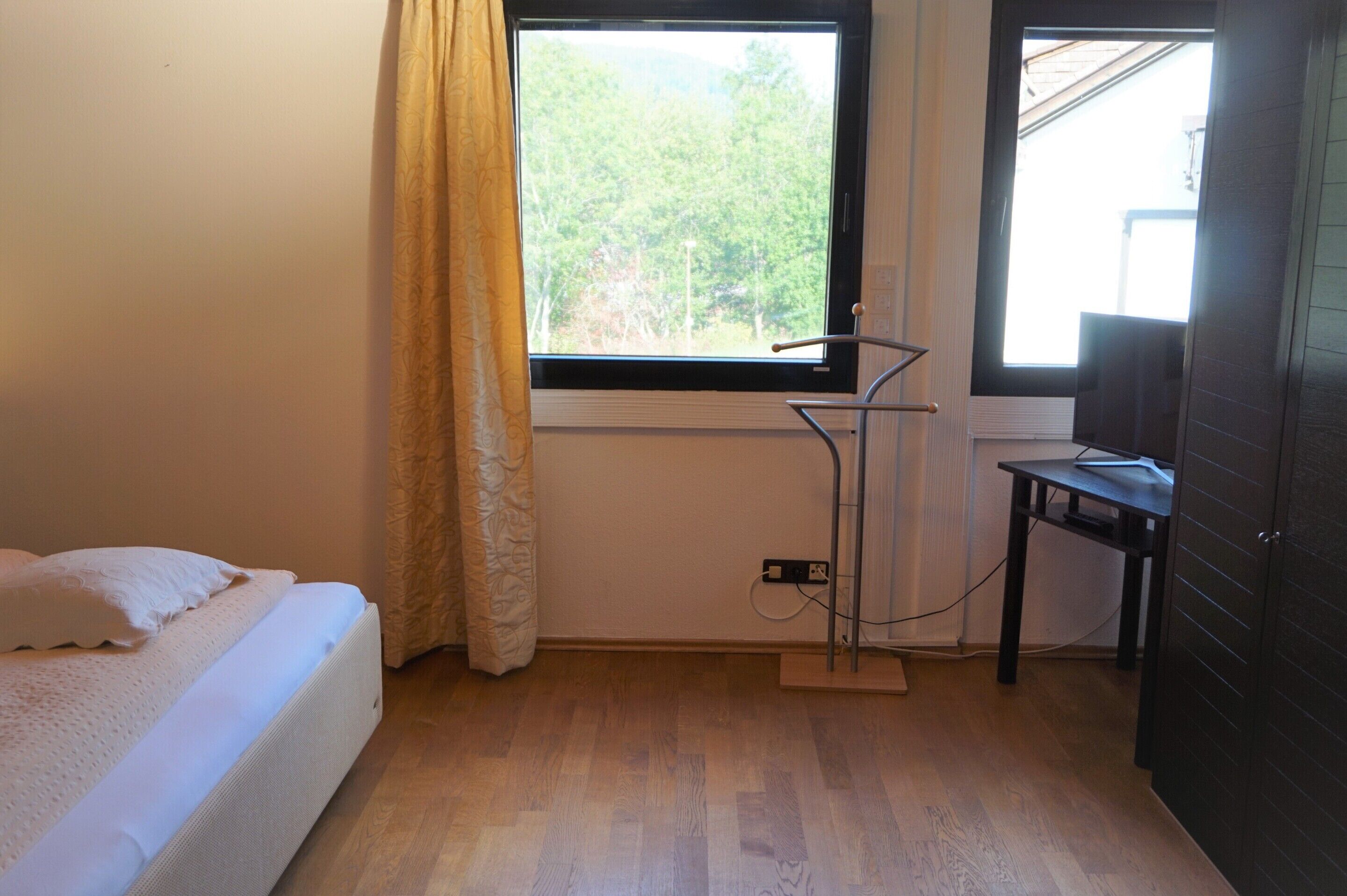 Un dormitorio con cama, ventana con vistas a la vegetación, radiador y escritorio con silla.