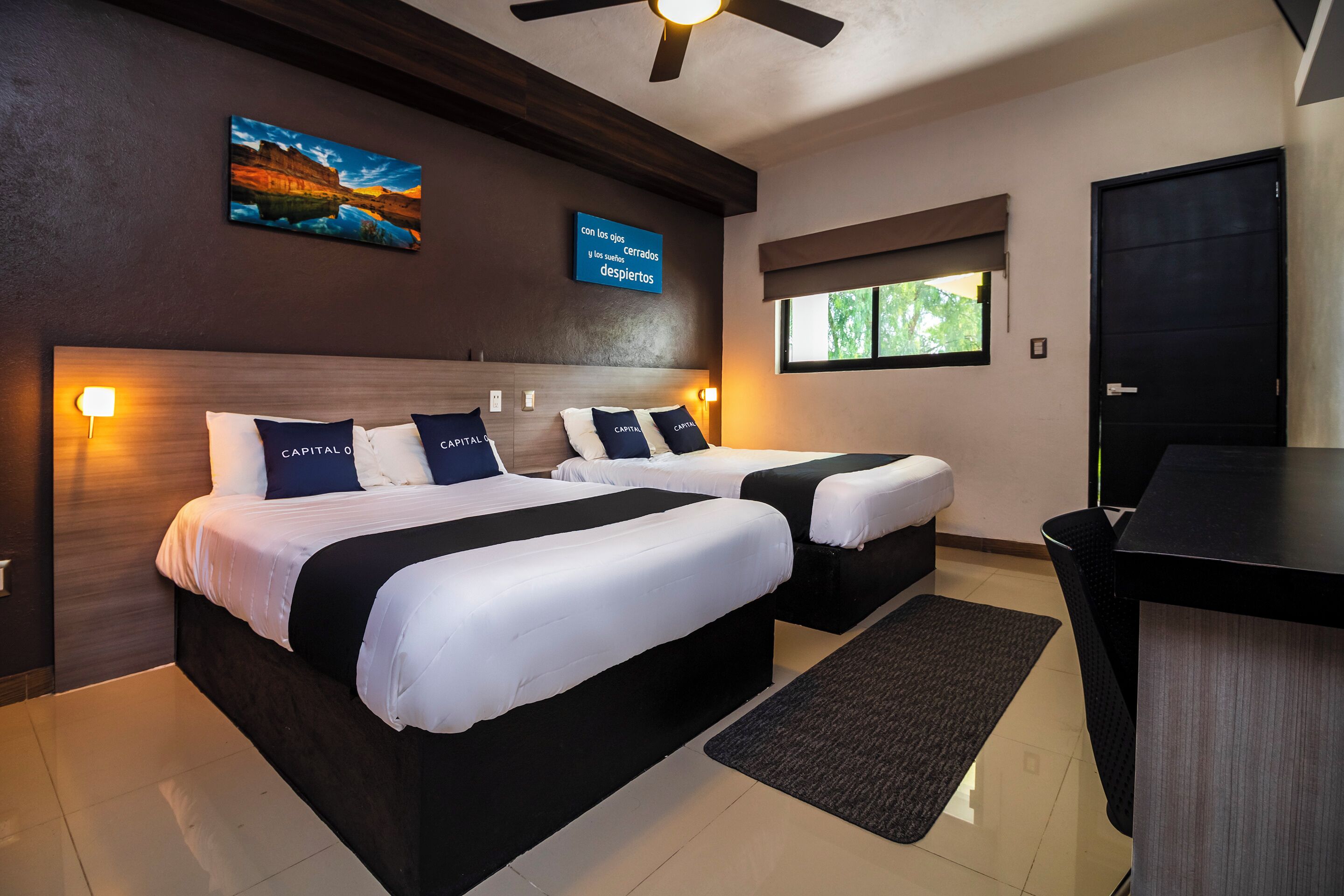 A hotel room with two beds, a desk, a ceiling fan, a window with a view, and a wall-mounted TV displaying text.