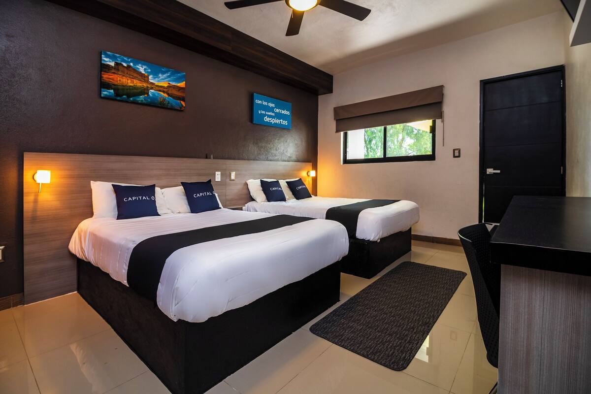 Habitación de hotel con dos camas, un escritorio, un ventilador de techo, una ventana con vistas y un televisor montado en la pared que muestra texto.