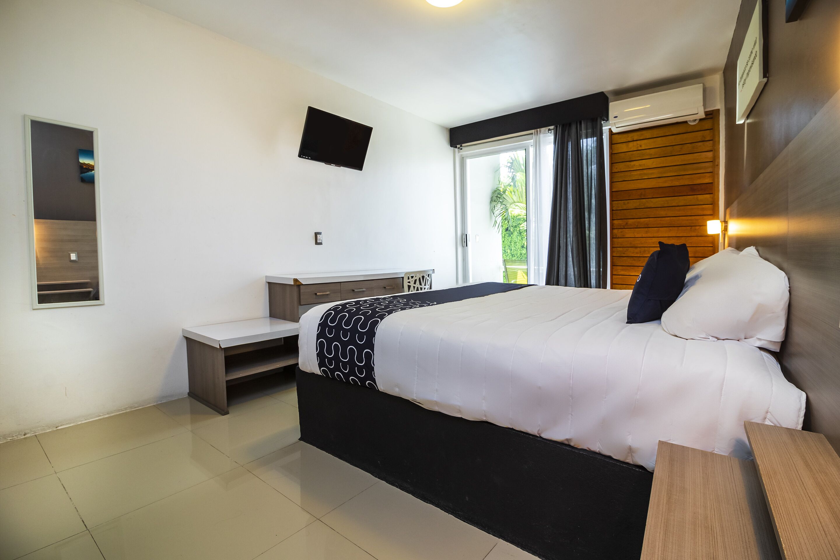 A modern hotel room with a large bed, a desk, a flat-screen TV, and a window with a view of greenery.
