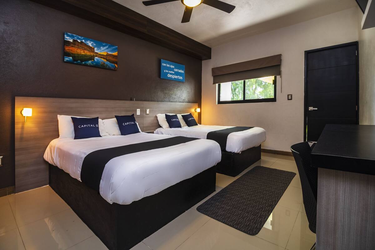 Habitación de hotel con dos camas, un escritorio, un ventilador de techo, una ventana con vistas y un televisor montado en la pared que muestra texto.