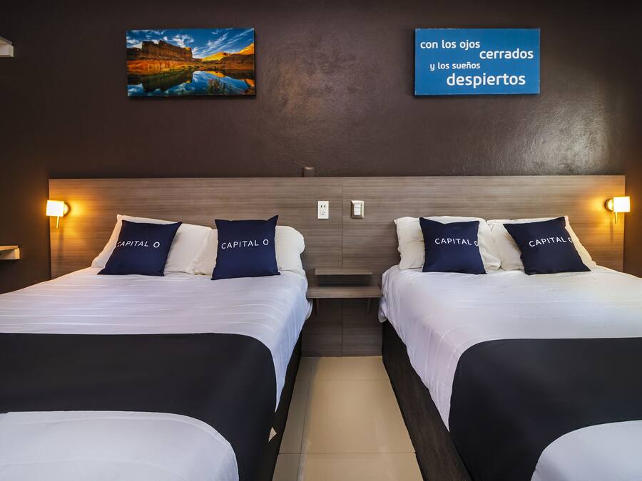 A hotel room with two beds, each with a white and black bedspread, and matching pillows. There are wall-mounted lamps on either side of the beds, and a painting above the beds.