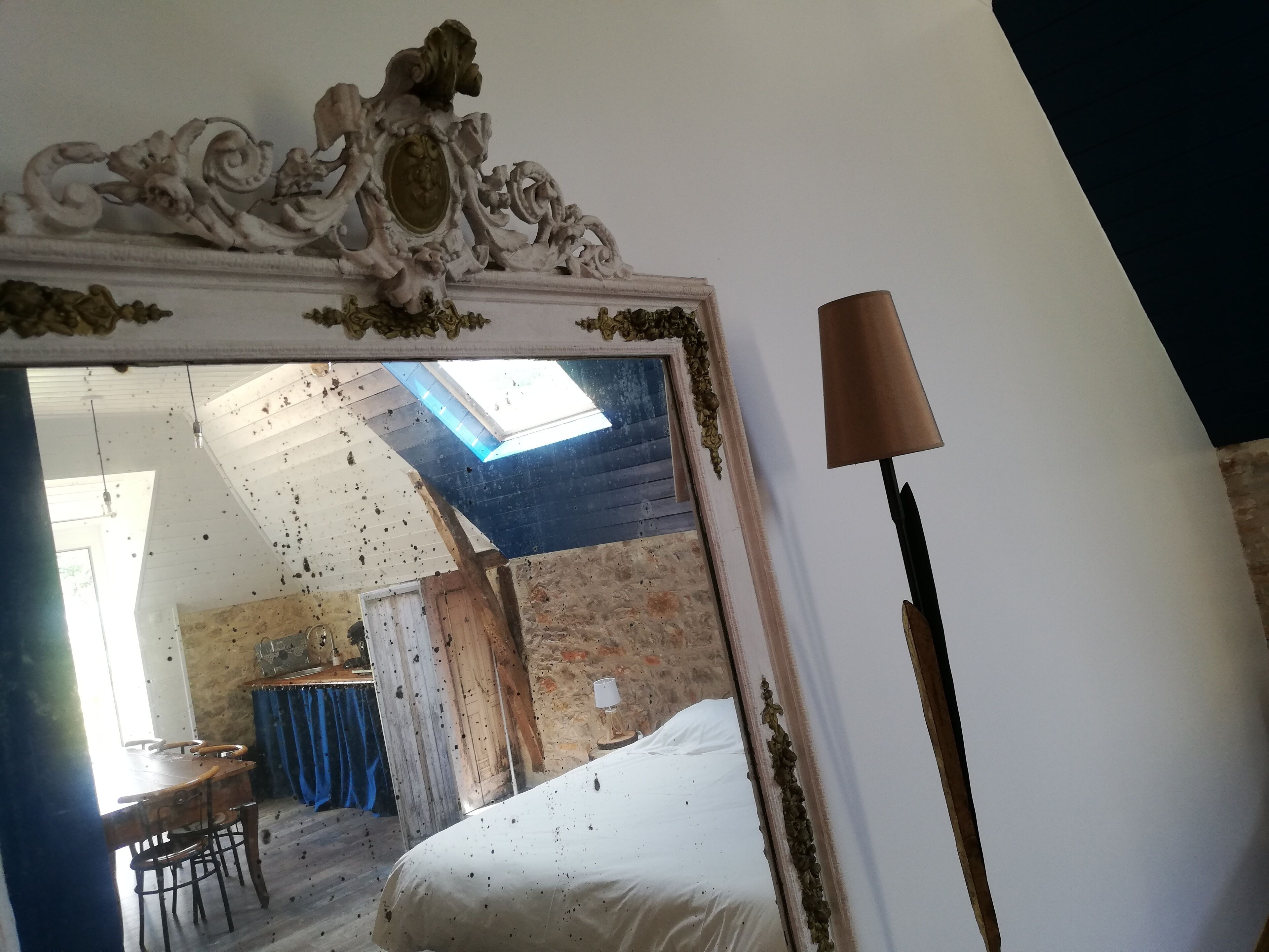 A room with a bed, a table, and a mirror reflecting a room with a blue ceiling and a window.
