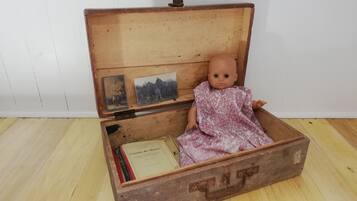 A vintage doll in a wooden box with a hinged lid, placed on a wooden floor.