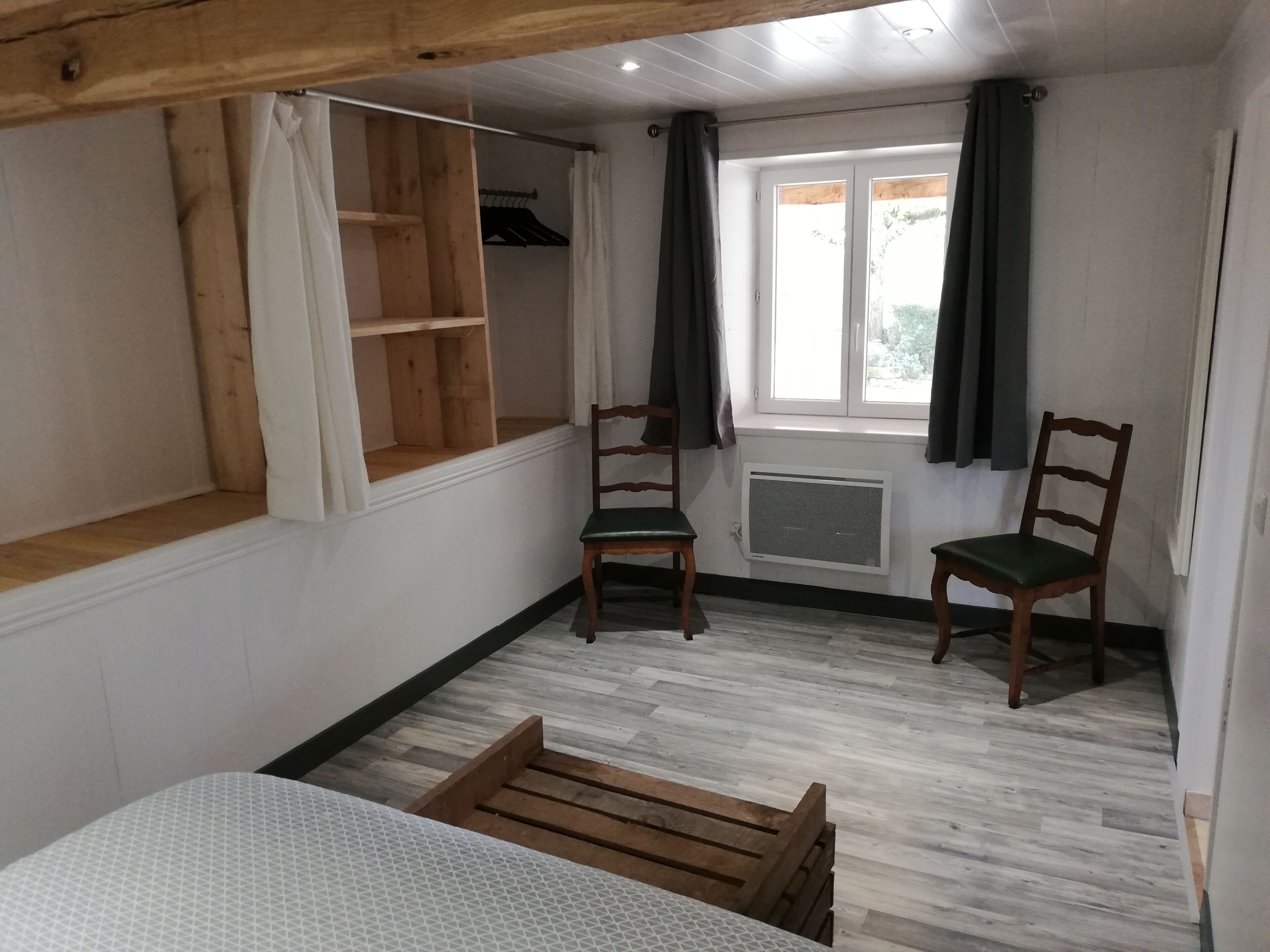 Una habitación con una cama, dos sillas de madera, una ventana con cortinas, un radiador y un estante de madera.