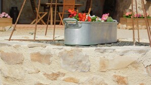 A rustic outdoor seating area with a metal pot containing flowers, a stone wall, and wooden furniture. - Le Puits de Garival (Tour-de-Faure)