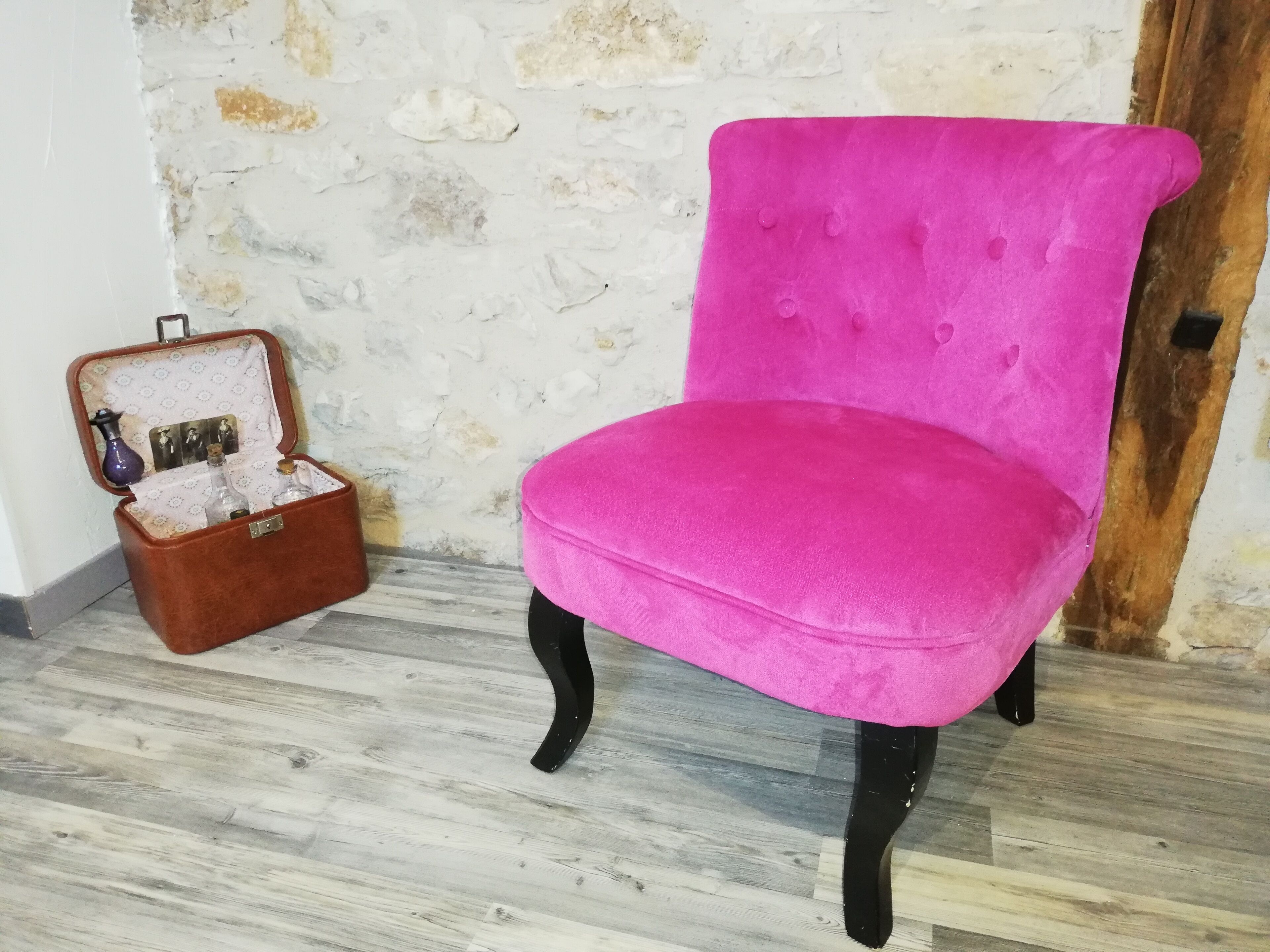 Una butaca rosa tapizada con patas negras, una maleta marrón con interior estampado, y una pared de piedra rústica.