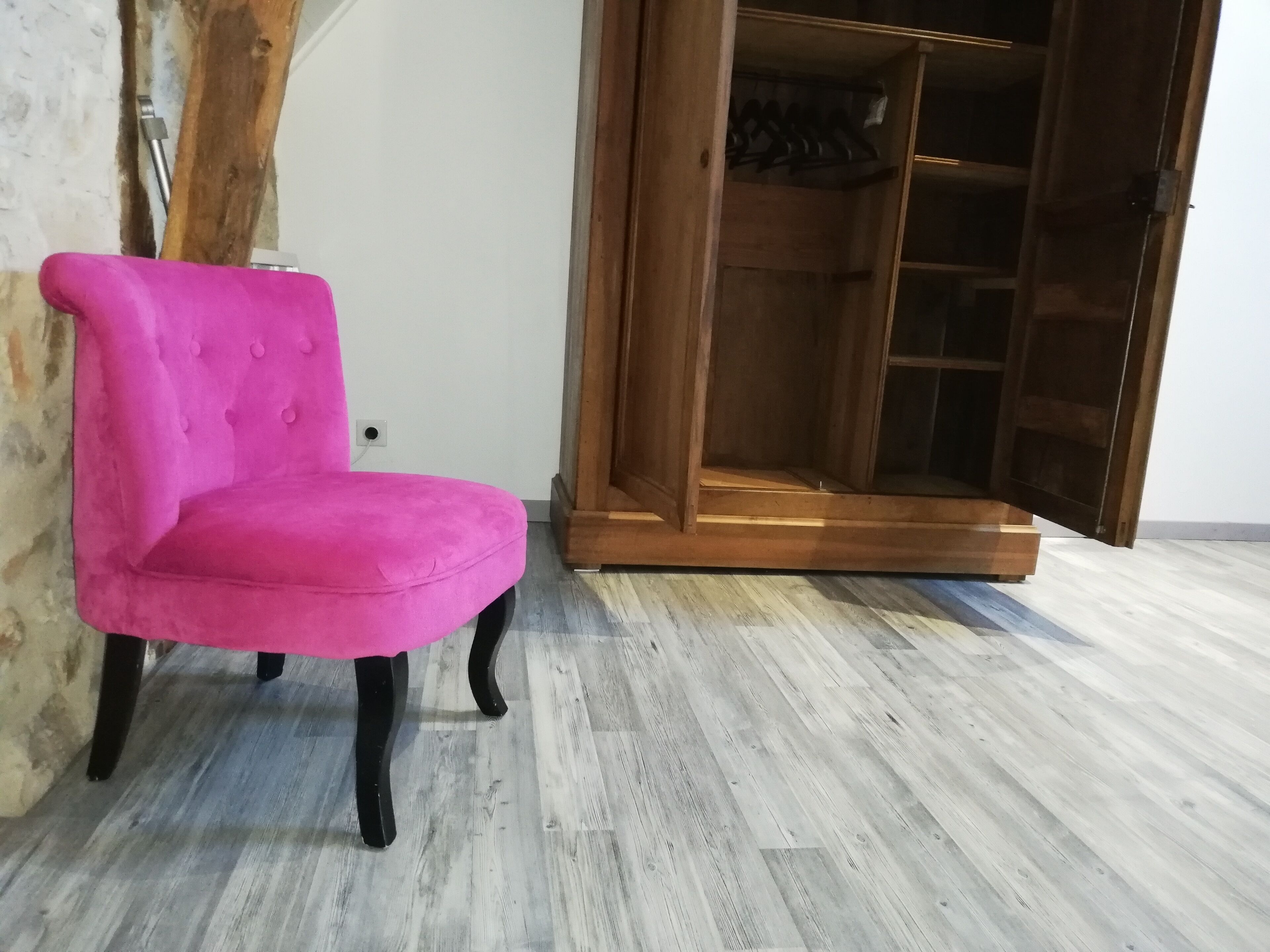 Una butaca rosa tapizada con patas negras en una habitación con suelo de madera y un armario de madera con puertas de vidrio.