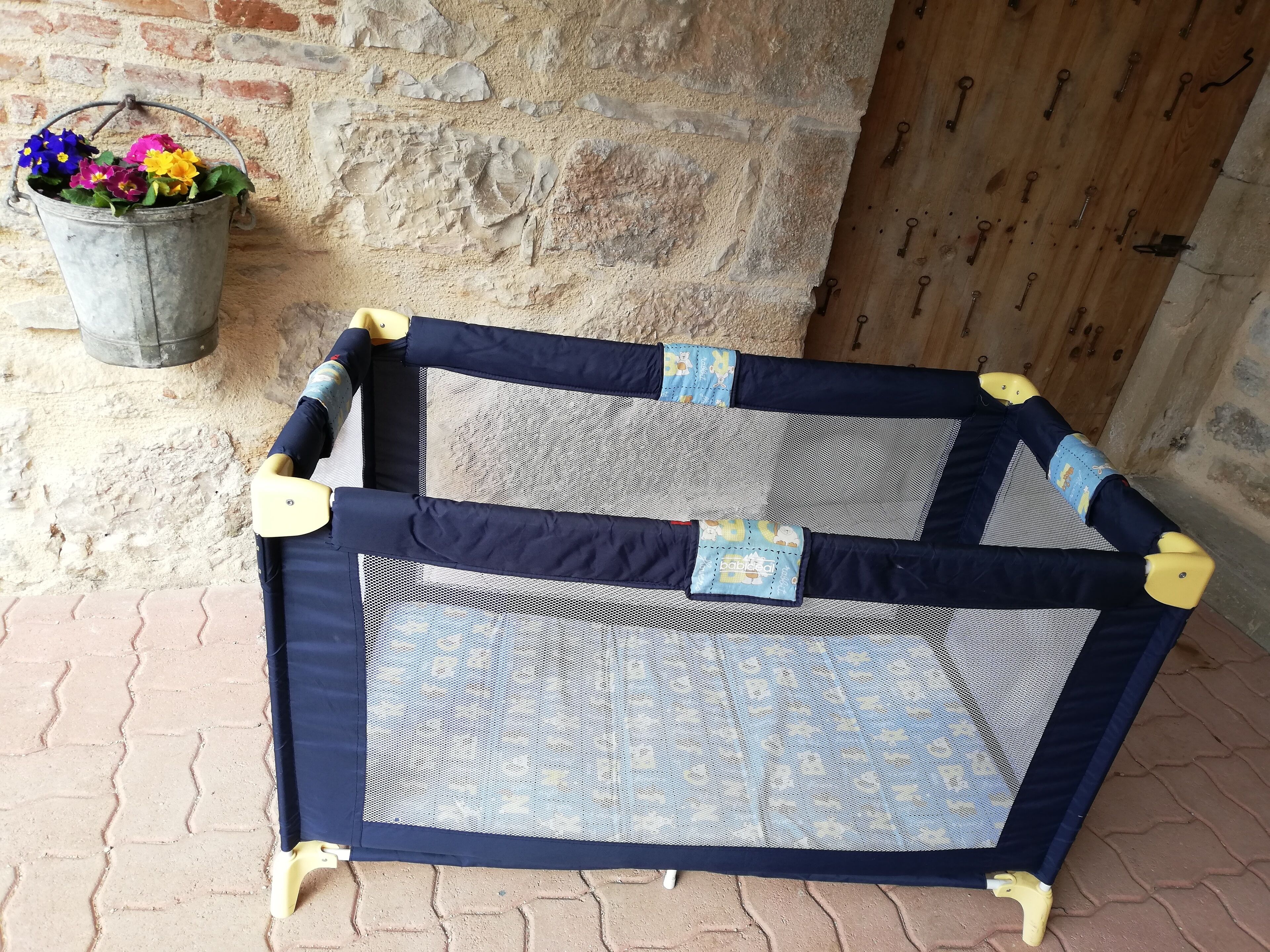 Een babyren met een gaaswand en een ontwerp in blauw en geel, geplaatst op een metselwerk vloer met een bakstenen muur en een houten deur op de achtergrond.