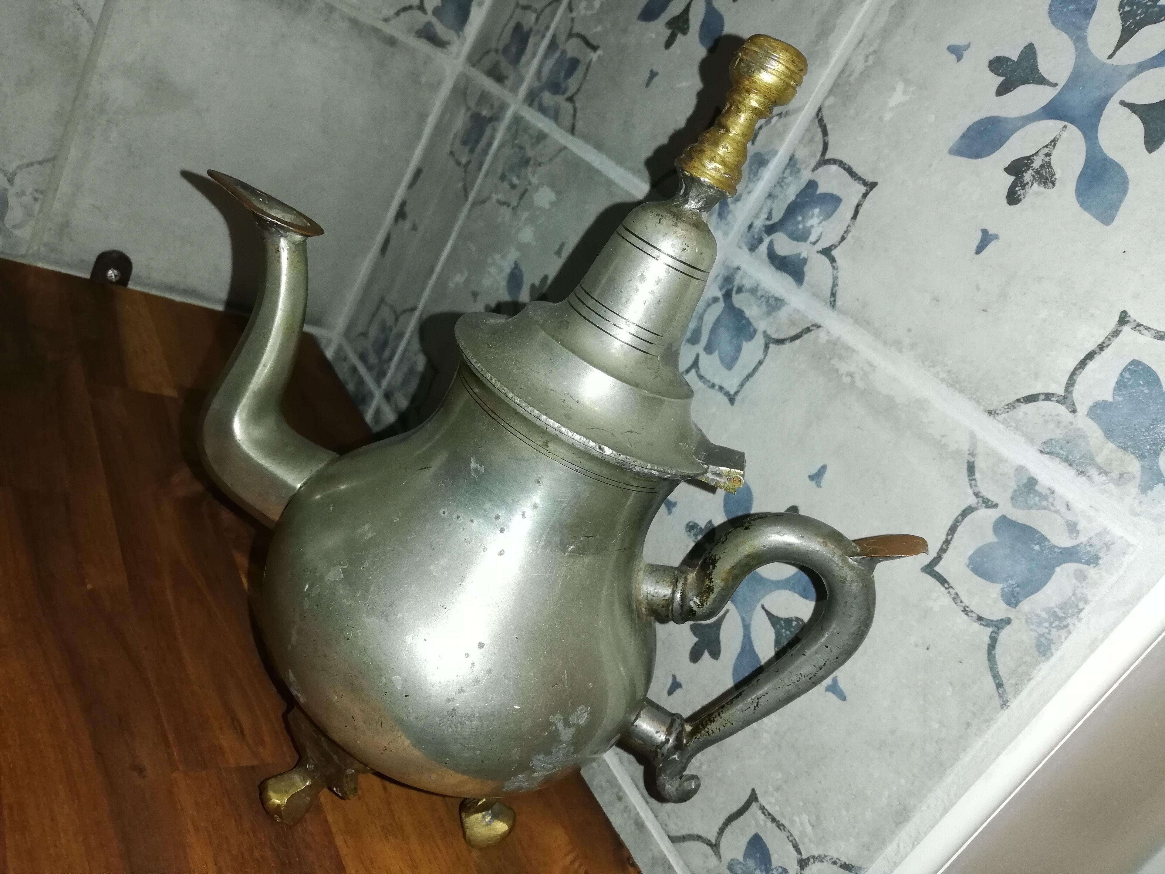 A metal teapot with a brass spout and handle, placed on a wooden surface against a tiled wall with a floral pattern.