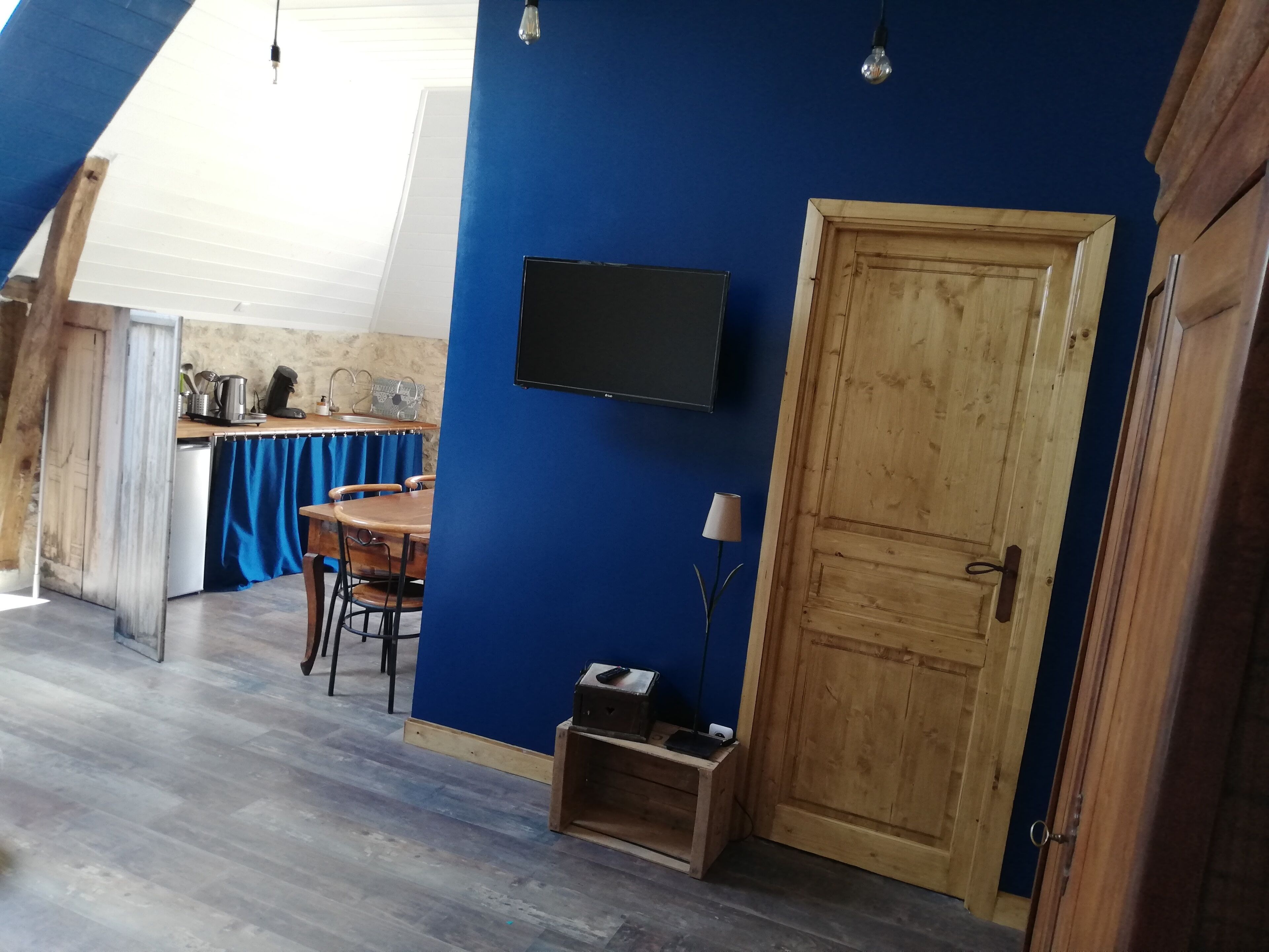 Una habitación con pared azul, puerta de madera, una pequeña mesa con sillas, televisor montado en la pared y una cocina al fondo.
