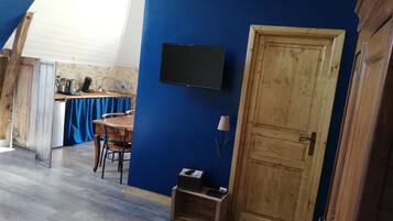 Ein Zimmer mit blauer Wand, einer Holztür, einem kleinen Tisch mit Stühlen, einem an der Wand befestigten Fernseher und einer Küchenzeile im Hintergrund.