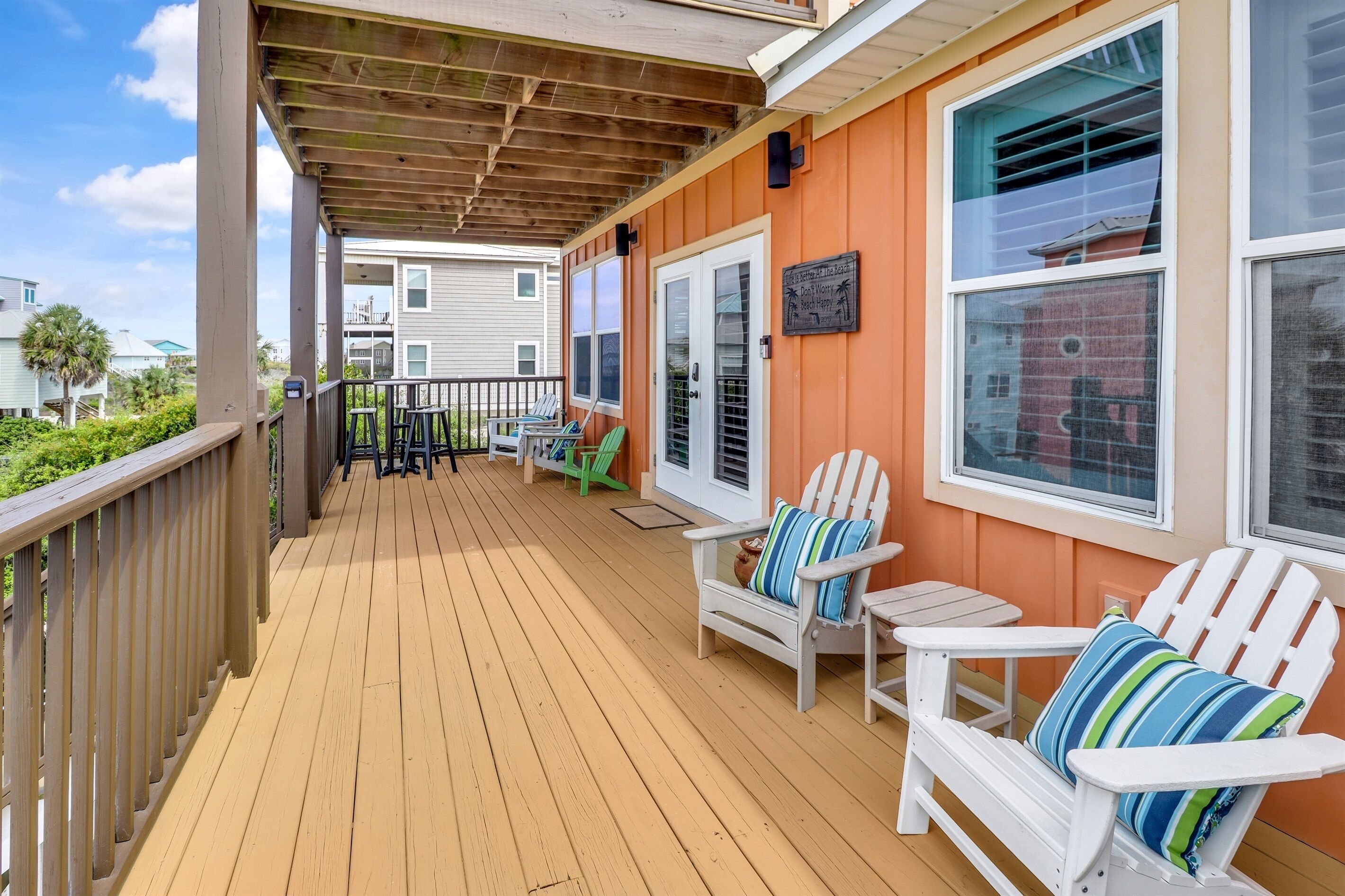 A wooden deck with white Adirondack chairs and a small table, a covered area with a pergola, and a view of neighboring buildings.