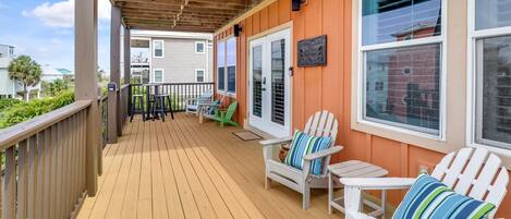 A wooden deck with white Adirondack chairs and a small table, a covered area with a pergola, and a view of neighboring buildings.